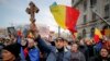 Демонстрант против вакцинација држи дрвен крст додека другите веат национални знамиња за време на протестите во Букурешт во март 2021 година.