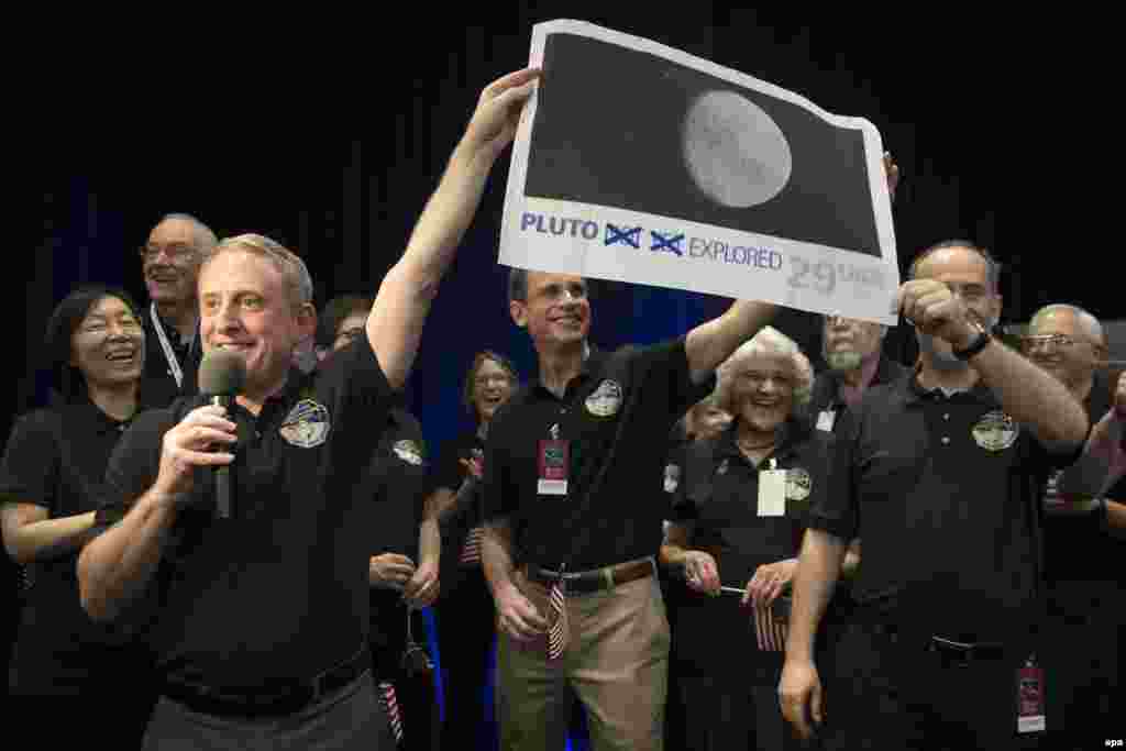 Ученые в центре контроля миссии New Horizons с увеличенным изображением 29-центовой почтовой марки, теперь устаревшей: на ней написано "Плутон еще не исследован" - на изображении слова "еще не" зачеркнуты. 
