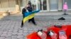 Жандармерист избутва фотографа Рафаело Казаков, който държи украинското знаме пред входа на руското консулство в София. На преден план - червена боя, която символизира кръвта на украински деца. В розово - манекен с фигура на момиче.