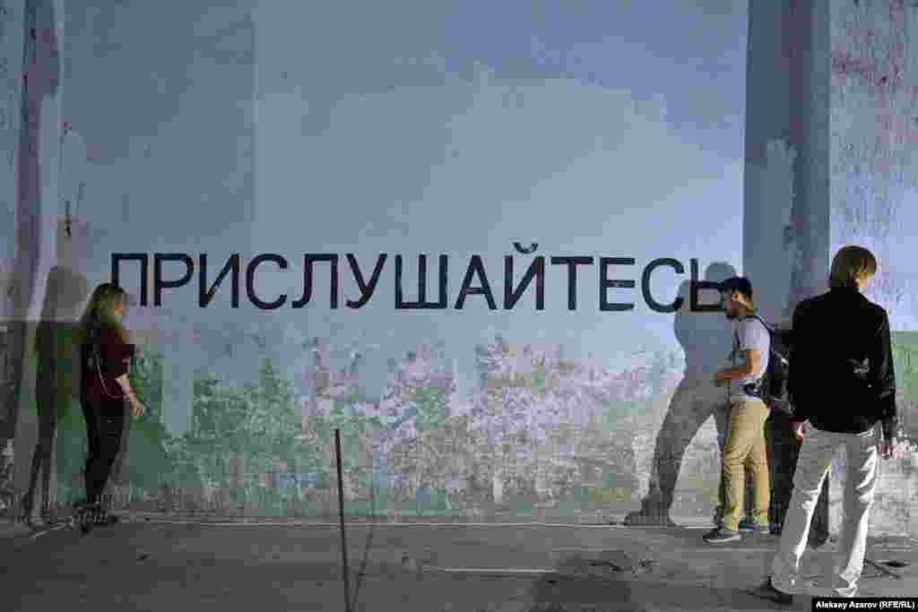 Девушка, обозначенная в каталоге выставки как Мира Евгеньевна, создала инсталляцию из надписи на стене: «Прислушайтесь», динамиков и аудиозаписей. Если приложить ухо к динамикам, то с одной стороны слышны сводки новостей нашего времени, а с другой стороны — записи выступлений деятелей партии «Алаш», «Алаш-Орды». Эта инсталляция — плод размышления о «деградации современного человека», о людях, не образованных как умственно, так и эстетически. 